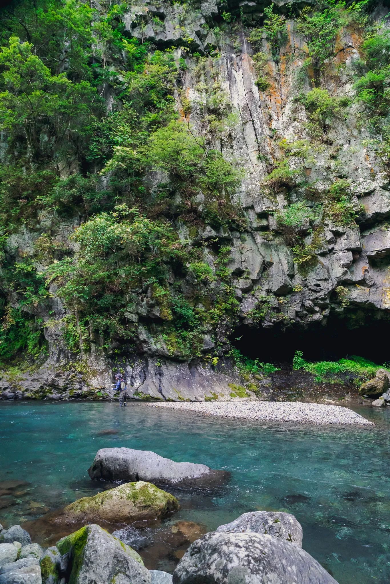 An angler small against a mossy cliff above turquoise water
